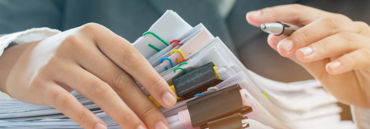 Mani di un professionista che esaminano una alta pila di documenti organizzati con clip colorate e penna in mano