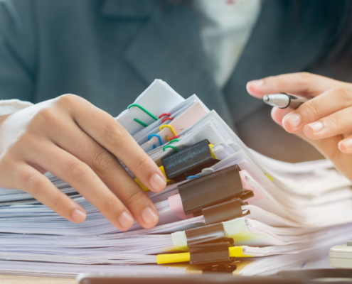Mani di un professionista che esaminano una alta pila di documenti organizzati con clip colorate e penna in mano