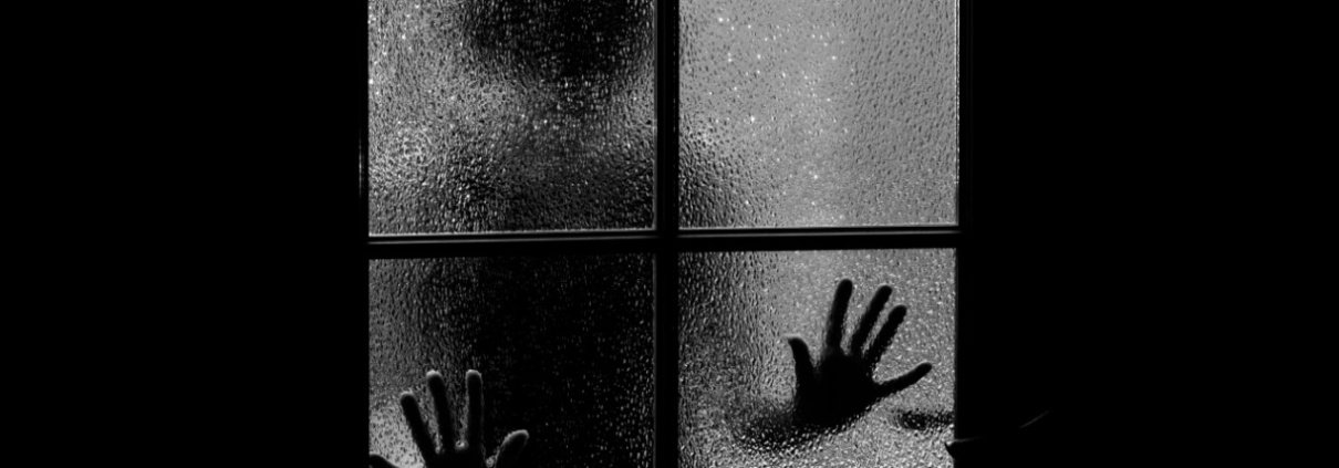 Silhouette scura di una persona con le mani appoggiate al vetro appannato di una finestra a quattro riquadri