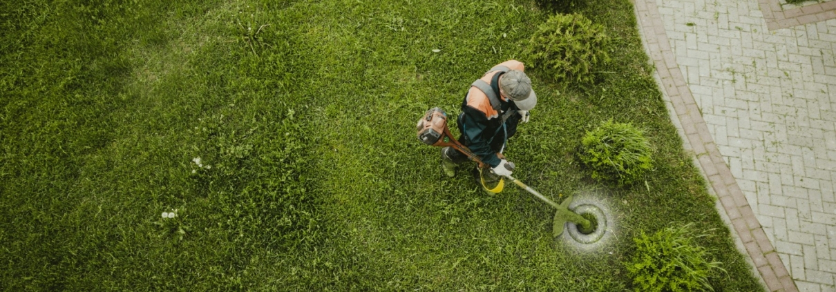 Vista dall'alto di un operaio con decespugliatore che cura un prato verde lungo un vialetto in mattoni
