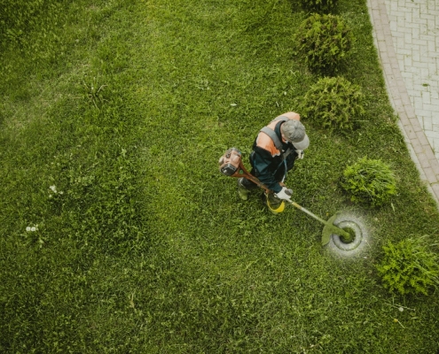 Vista dall'alto di un operaio con decespugliatore che cura un prato verde lungo un vialetto in mattoni