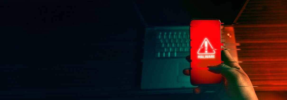 Mano che tiene uno smartphone con schermo rosso e avviso malware, accanto a un portatile in penombra